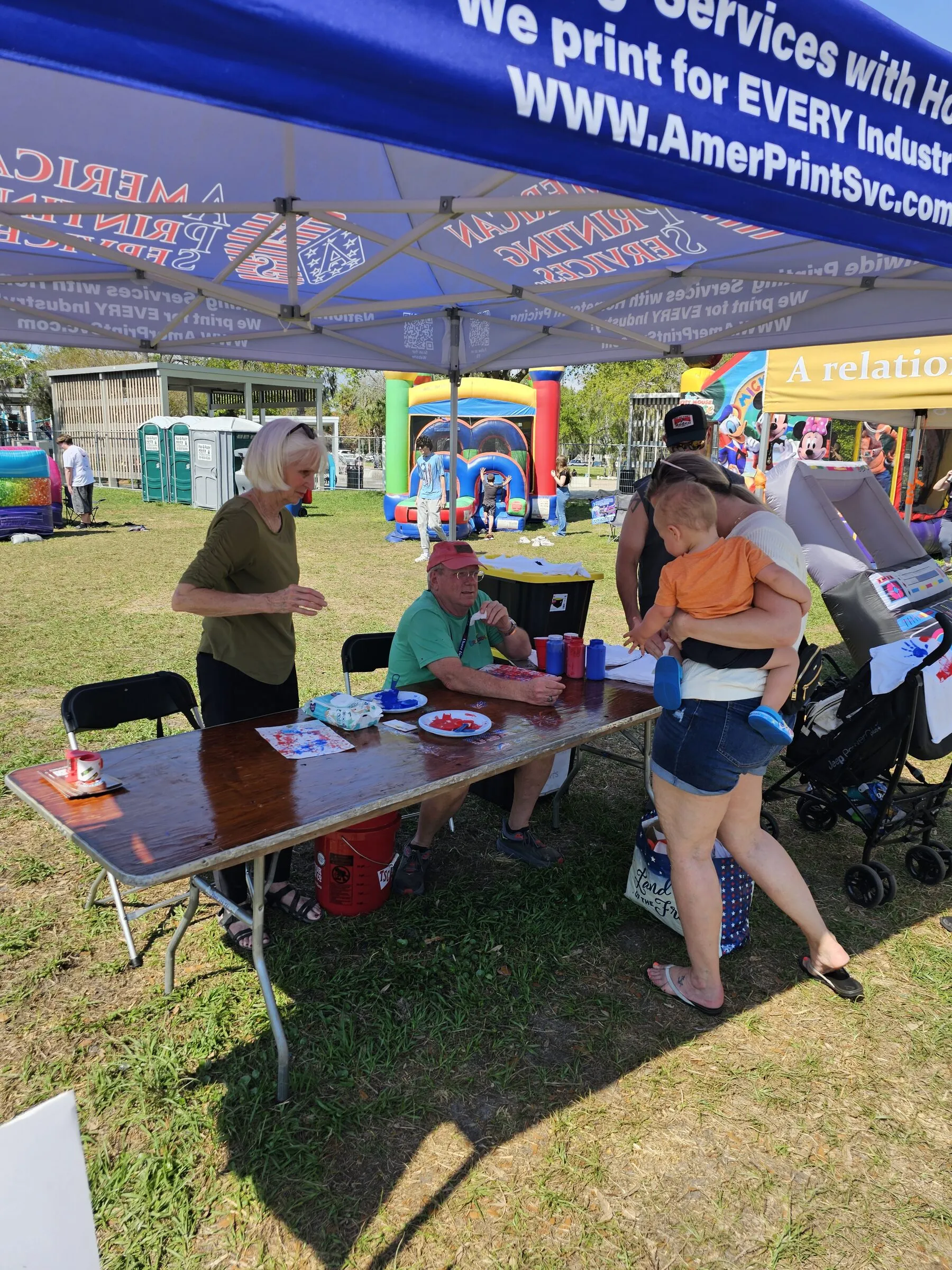 Families gather at an activity table under a sponsor tent at an outdoor community festival, with bounce houses in the background