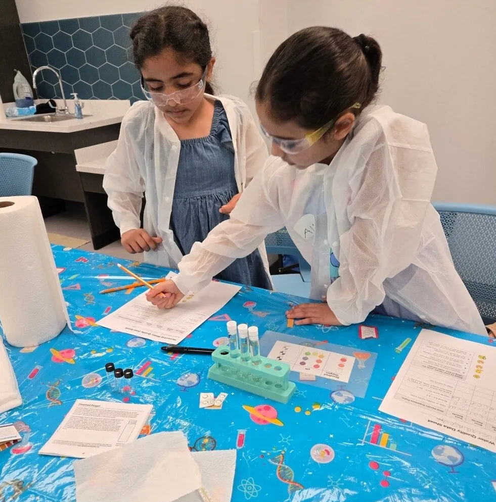 Two students in lab coats lean over a science activity at a Science Saturday table
