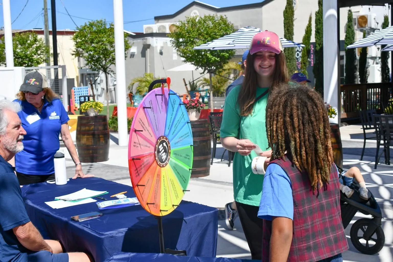 Brainstorms volunteers greet a visitor at an outdoor popup table with a colorful prize wheel